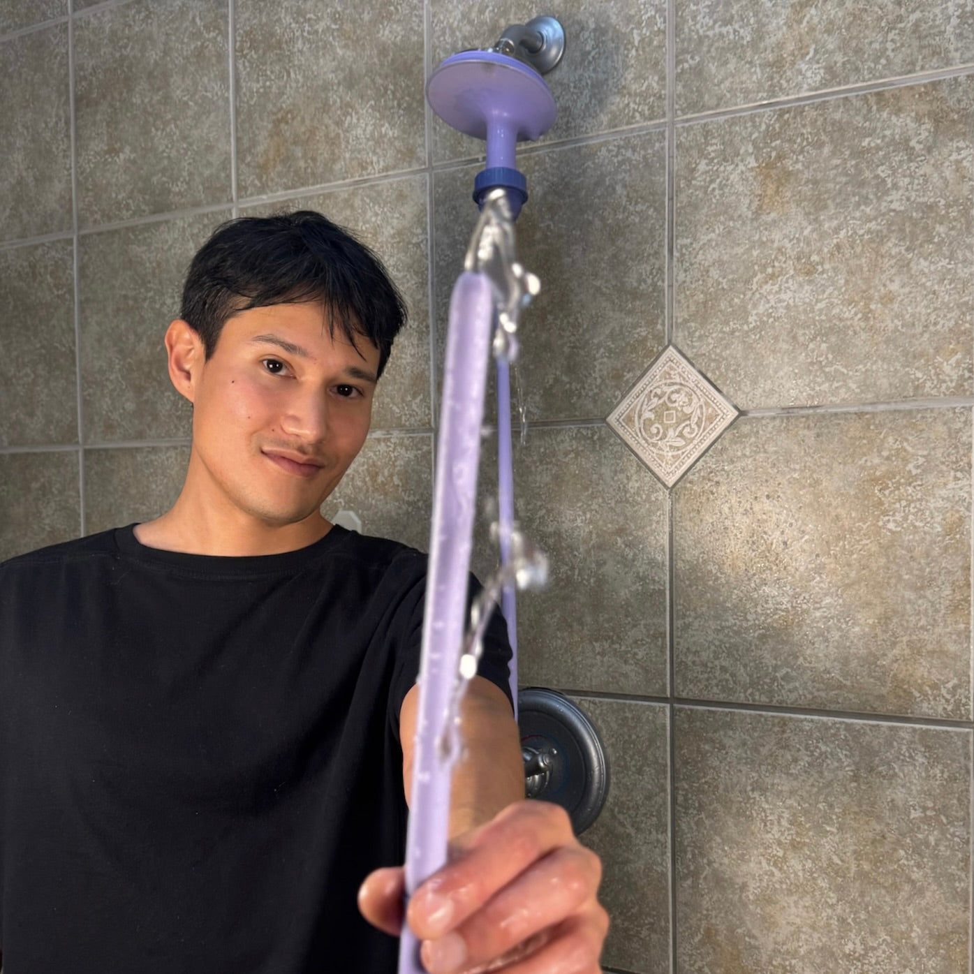 Person holding Squwash shower enema douche attached to shower head against a tiled bathroom wall as water flows through the tip