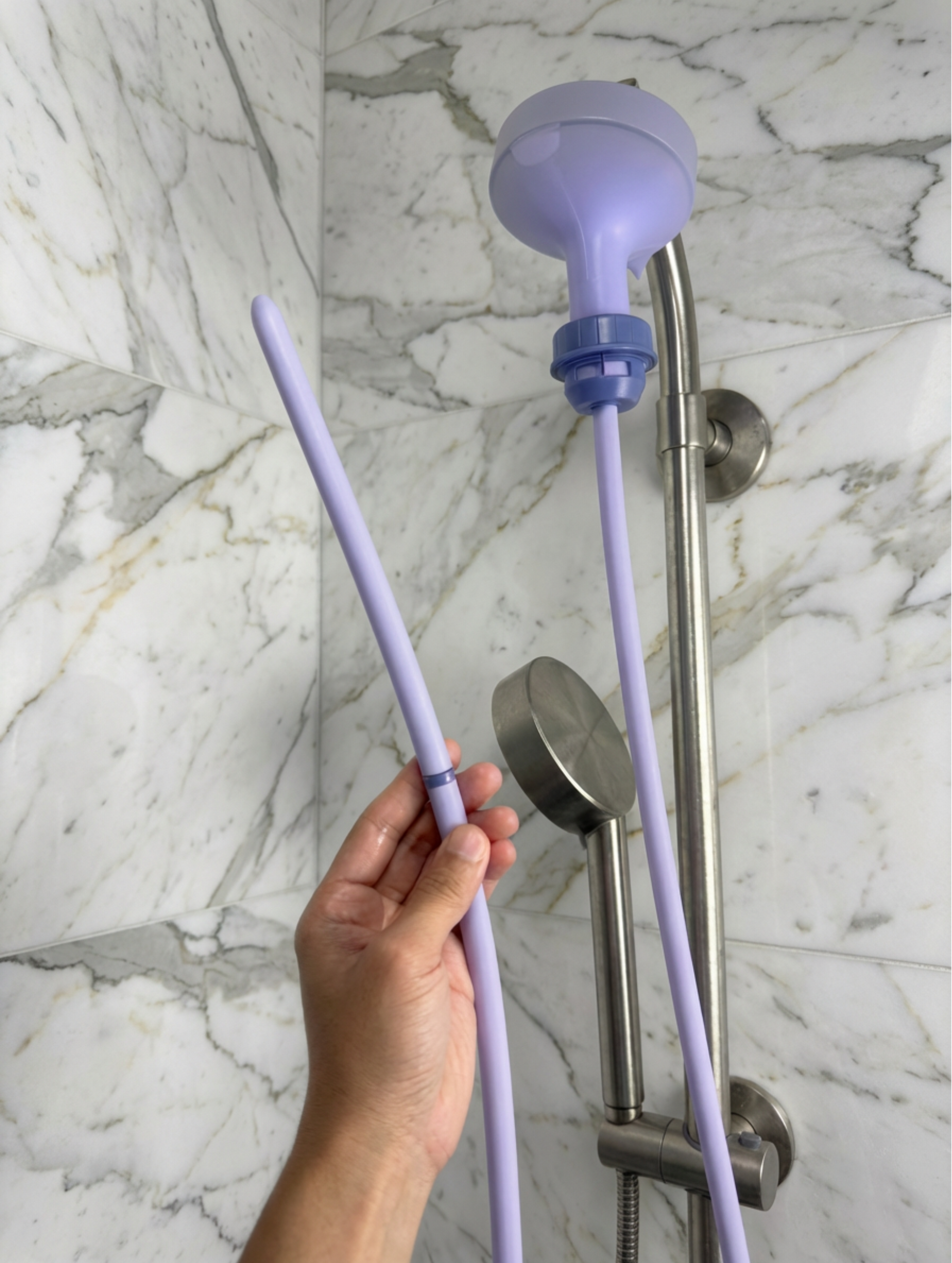 Purple Squwash shower head extension held by a hand against a marble shower wall.