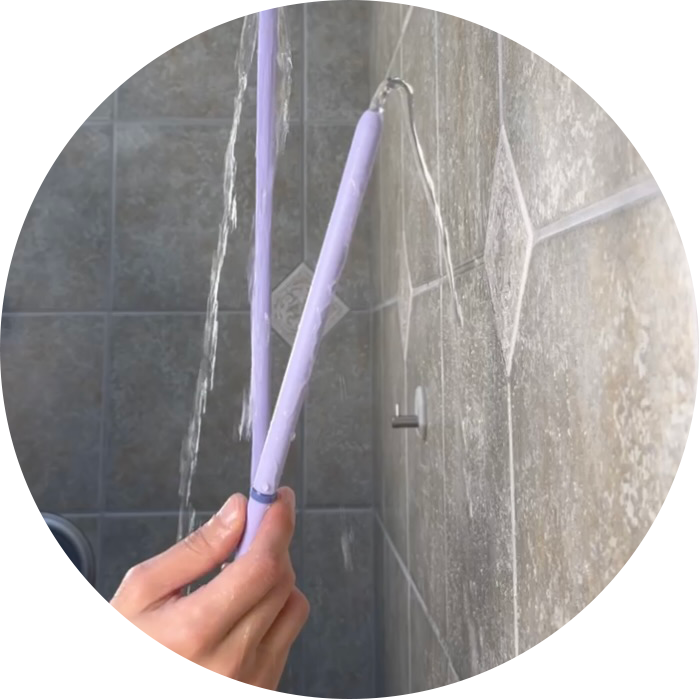 Hand holding purple platinum-cured silicone tip of 'Squwash' branded portable shower douche against a tiled shower wall as water flows through tip.