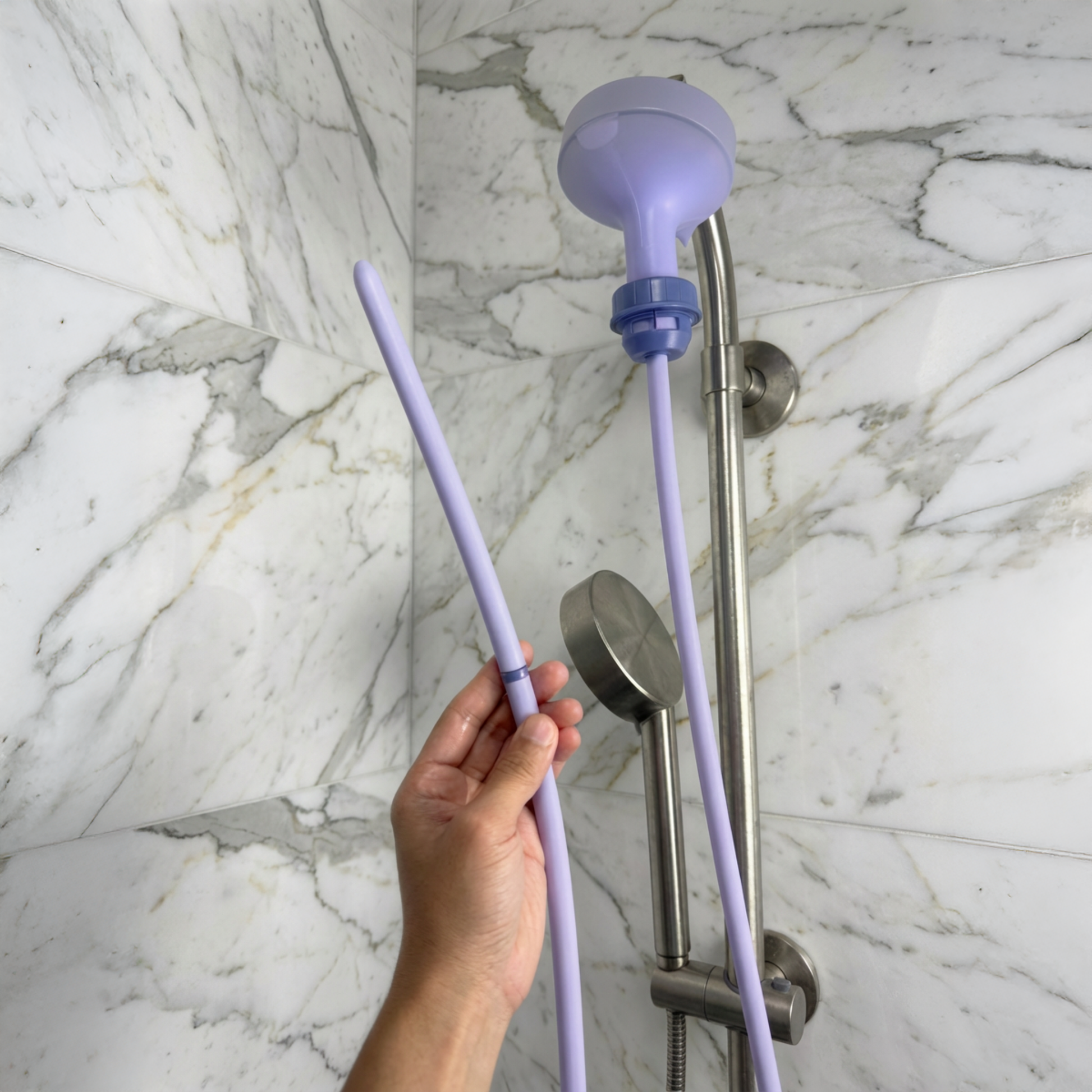 Purple Squwash slip-on enema douche shower head extension held by a hand against a marble shower wall.
