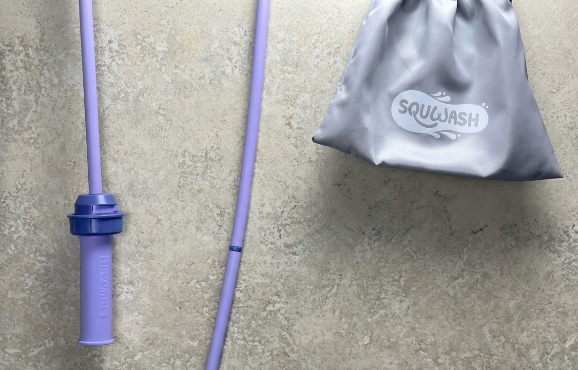 Purple Squwash hose hanging in bathroom against a concrete surface, next to a gray drawstring travel bag with 'Squwash' branding.