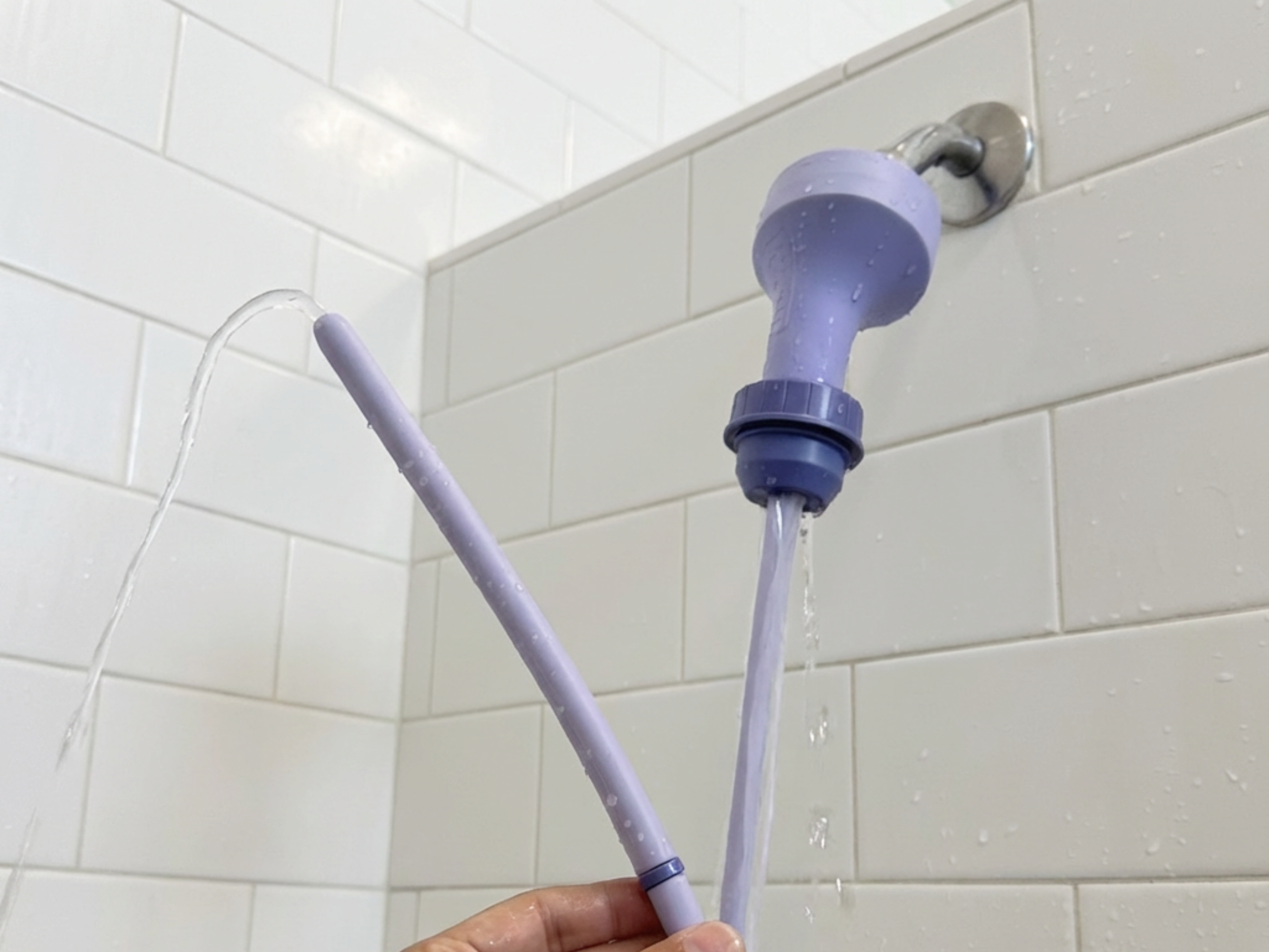 Purple Squwash shower hygiene tool travel douche enema kit with silicone hose and tip in gym shower as water is flowing through the device shown against a white tile bathroom wall 